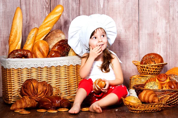 头上有面包。戴着厨师帽的面带微笑的小女孩在一个柳条篮子附近吃面包和百吉饼, 里面有面包卷和面包烘焙产品 — 图库照片