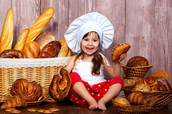 Chléb, kolem hlavy. Usměvavá holčička v čepici kuchař jí chléb a rohlíky poblíž proutěný koš s pečivo a pekárenské výrobky — Stock fotografie