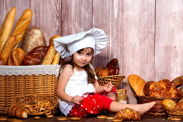 Pão à volta da cabeça. Pequena menina sorridente em um tampão de cozinha come pão e bagels perto de uma cesta de vime com pães e produtos de padaria — Fotografia de Stock