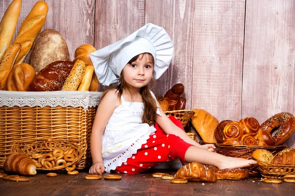 头上有面包。戴着厨师帽的面带微笑的小女孩在一个柳条篮子附近吃面包和百吉饼, 里面有面包卷和面包烘焙产品 — 图库照片