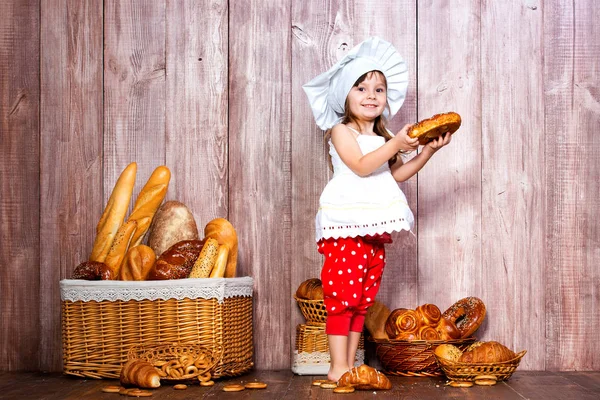 Essen frisches Brot und hausgemachtes Gebäck. kleines lächelndes Mädchen in Kochmütze mit Bagel in der Hand neben einem Weidenkorb mit Brötchen und Backwaren — Stockfoto