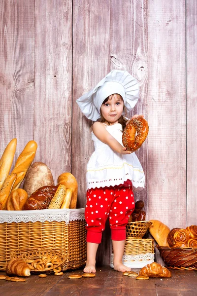 Eet vers brood en huisgemaakt gebak. Beetje glimlachen meisje in een cook dop met bagel in de hand in de buurt van een rieten mand met broodjes en bakkerijproducten — Stockfoto