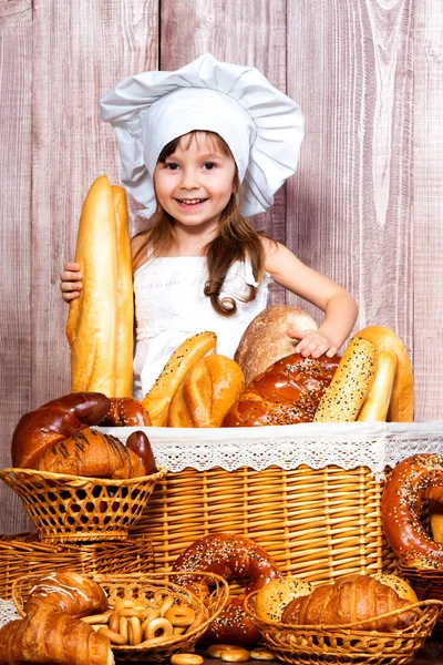 Süßes lächelndes Mädchen mit Kochmütze, gebacken in der Nähe eines Weidenkorbs mit frischen Brötchen, hausgemachtem Gebäck und Backwaren — Stockfoto
