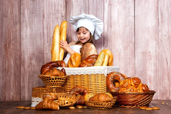 Милая улыбающаяся девушка в шапке повара, запечённая возле плетеной корзины со свежими булочками, домашней выпечкой и хлебобулочными изделиями — стоковое фото