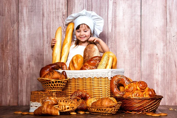 Menina sorridente bonito em um tampão de cozinheiro assado perto de uma cesta de vime com pães frescos, doces caseiros e produtos de padaria — Fotografia de Stock