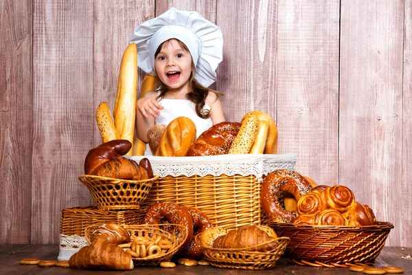 Menina sorridente bonito em um tampão de cozinheiro assado perto de uma cesta de vime com pães frescos, doces caseiros e produtos de padaria — Fotografia de Stock