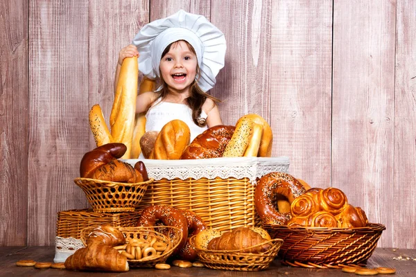 Menina sorridente bonito em um tampão de cozinheiro assado perto de uma cesta de vime com pães frescos, doces caseiros e produtos de padaria — Fotografia de Stock