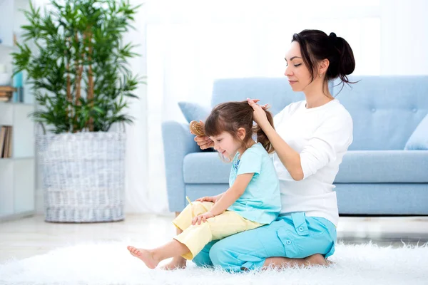 Счастливая семья. Мама расчесывает дочери волосы, сидя на ковре на полу в комнате. — стоковое фото