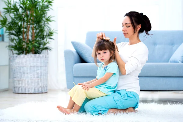 Feliz família amorosa. Mãe está penteando o cabelo de sua filha sentado no tapete no chão na sala — Fotografia de Stock