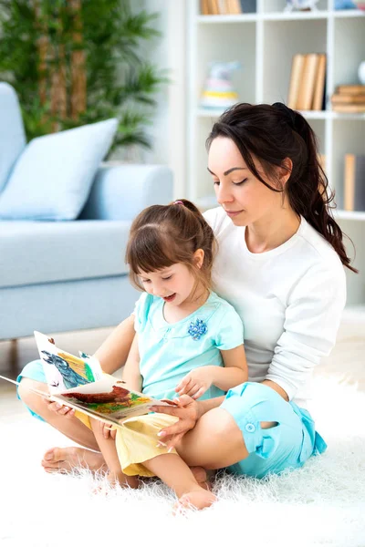 Bela jovem mãe lendo um livro para sua filha sentada no tapete no chão da sala. Leitura com crianças — Fotografia de Stock