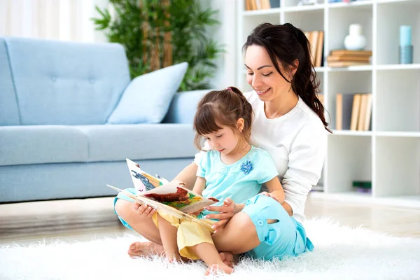 Досить молода мати читає книжку доньці, сидячи на килимі на підлозі в кімнаті. Читання з дітьми — стокове фото
