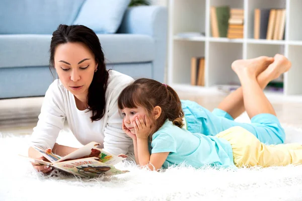 Досить молода мати читає книгу свою маленьку доньку, лежачи на килимі на підлозі вдома — стокове фото