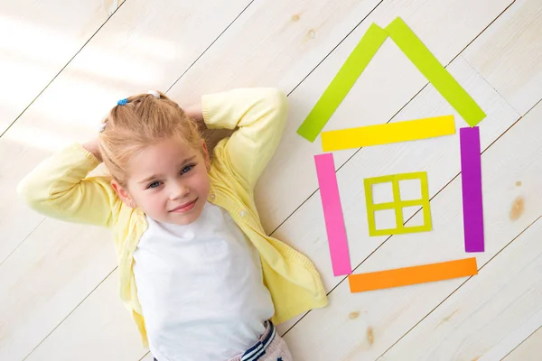 Маленькая девочка лежит на полу рядом с цветным бумажным домом — стоковое фото
