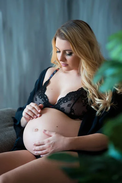 Těhotná fotofotografie v podkroví. Krásná těhotná matka se světlými vlasy sedí na pohovce a zasněně se dívá z okna a na břicho — Stock fotografie