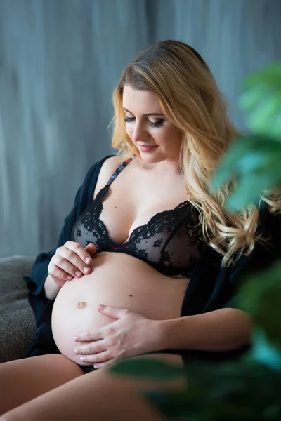 Pregnant photoshoot in loft style. Beautiful pregnant mother with blond hair sits on sofa and dreamily looks out the window and on tummy — Stock Photo, Image