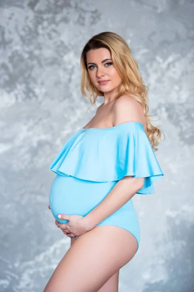 Vacker ung gravid blond kvinna i en blå kostym kropp mot en grå betongvägg bakgrund. Kvinnors hälsa och vackra graviditet — Stockfoto
