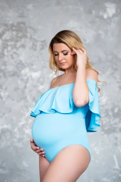 Mooie jonge zwangere blonde vrouw in een blauw pak lichaam tegen een grijze betonnen muur achtergrond. Vrouwen gezondheid en mooie zwangerschap — Stockfoto
