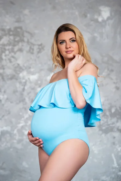 Bela jovem mulher loira grávida em um corpo de terno azul contra um fundo de parede de concreto cinza. Saúde da mulher e bela gravidez — Fotografia de Stock