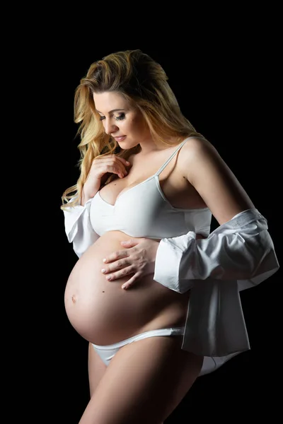 Mooie blonde zwangere vrouw in witte kleren op een zwarte achtergrond. Vrouwen gezondheid en gelukkige zwangerschap — Stockfoto