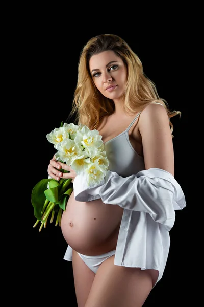 Krásná blondýnka s těhotným nádmečkem na bílém tričku a držící v rukou květiny bílé tulipány. Dívka pózní na černém pozadí — Stock fotografie