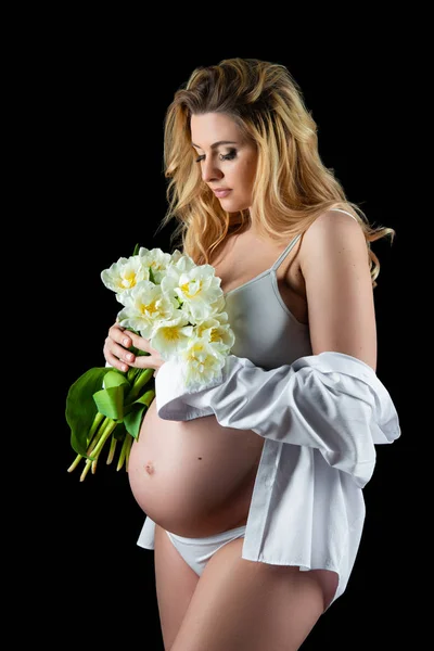美丽的金发女子与怀孕的肚子穿着白色衬衫，手里拿着鲜花白色的郁金香。女孩在黑色背景上摆姿势 — 图库照片