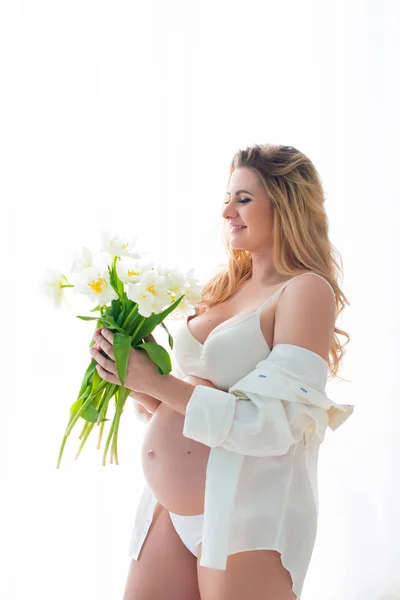 Femeie gravidă frumoasă cu flori albe de primăvară în fața unei ferestre inundate de lumina soarelui. Sănătatea femeii și sarcina fericită — Fotografie, imagine de stoc