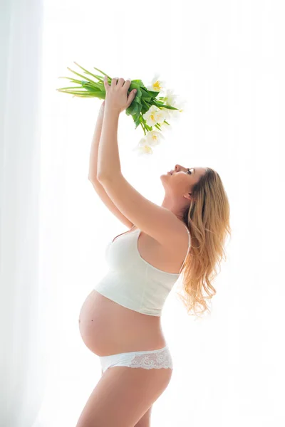 Mooie zwangere vrouw met lente witte bloemen voor een venster overspoeld met zonlicht. Vrouwelijke gezondheid en gelukkige zwangerschap — Stockfoto