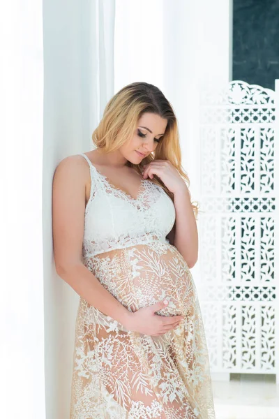 Een mooie zwangere vrouw in mooie kanten jurk staat in de buurt van een helder raam. Vrouwelijke seksualiteit, vrouwelijkheid en mooie zwangerschap — Stockfoto