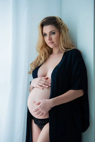Uma bela mulher grávida em um manto preto fica perto de uma janela brilhante. Sexualidade feminina, feminilidade e bela gravidez — Fotografia de Stock