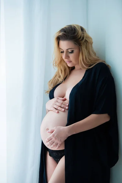 A beautiful pregnant woman in a black robe stands near a bright window. Feminine sexuality, femininity and beautiful pregnancy — Stock Photo, Image