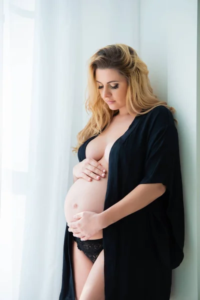 Een mooie zwangere vrouw in een zwarte gewaad staat in de buurt van een helder raam. Vrouwelijke seksualiteit, vrouwelijkheid en mooie zwangerschap — Stockfoto