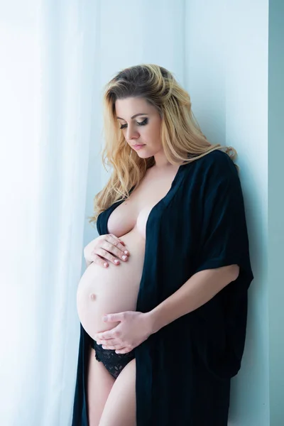 En vacker gravid kvinna i en svart mantel står nära ett ljust fönster. Kvinnlig sexualitet, kvinnlighet och vacker graviditet — Stockfoto