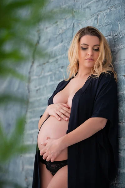 Uma bela mulher grávida em um manto preto se ergue contra a parede de tijolo cinza. Sexualidade feminina, feminilidade e bela gravidez — Fotografia de Stock
