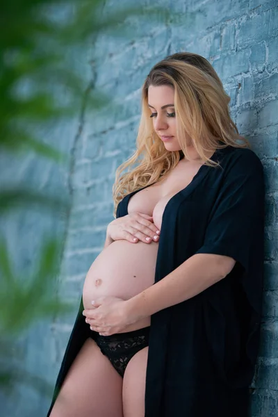 A beautiful pregnant woman in a black robe stands on against the gray brick wall. Feminine sexuality, femininity and beautiful pregnancy — Stock Photo, Image