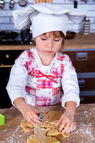 A girl in a chef's hat is cooking gingerbread in the kitchen, making cakes from cookies tins — Stock Photo, Image