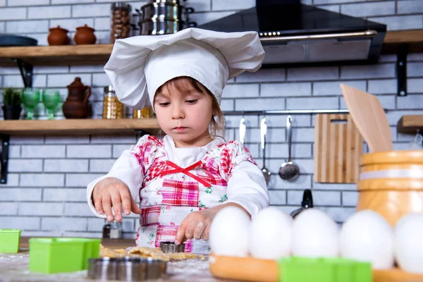 A girl in a chef's hat is cooking gingerbread in the kitchen, making cakes from cookies tins — Stock Photo, Image