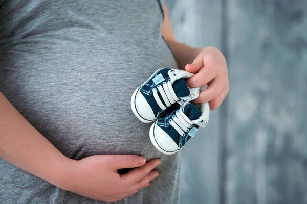 Boldog anya, megelőlegezve a fia születését. Terhes nő, aki Baby zsákmányt cipők a has háttérben. Közeli fel a terhes hasa — Stock Fotó