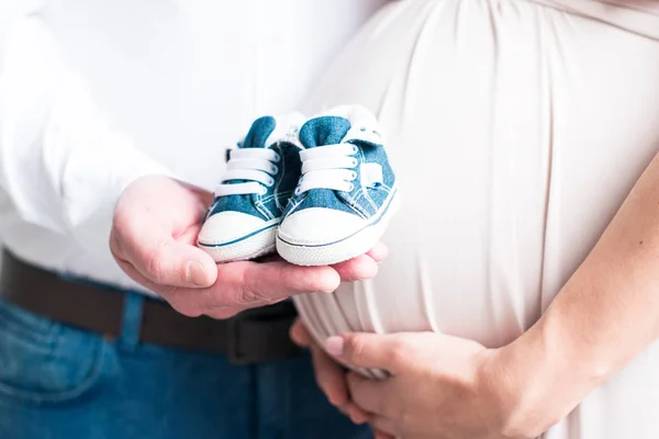 Närbild av en gravid mage. Lyckliga föräldrar väntar på sonen. Gravid par innehar baby tossor sneakers på magen bakgrund — Stockfoto