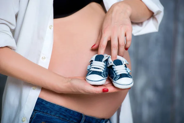 Boldog anya, megelőlegezve a fia születését. Terhes nő, aki Baby zsákmányt cipők a has háttérben. Közeli fel a terhes hasa — Stock Fotó