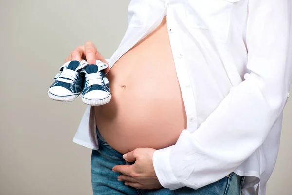 Feliz madre en anticipación del nacimiento de su hijo. Mujer embarazada sosteniendo botines de bebé zapatillas de deporte en el fondo de la panza. Primer plano de un vientre embarazada —  Fotos de Stock