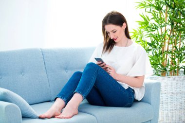 Genç bir kadın akıllı telefonuyla oynarken kanepede oturuyor.