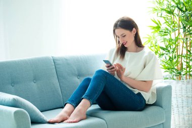 Genç bir kadın akıllı telefonuyla oynarken kanepede oturuyor.