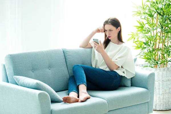 Young woman sitting on a sofa while playing with her smartphone — Stock Photo, Image