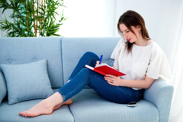 Student girl studing on a sofa with a red notebook. — Stock Photo, Image