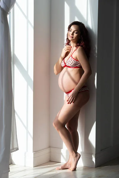 Piękna dziewczyna w ciąży w bieliźnie stoi w pobliżu okna. Młoda kobieta czeka na dziecko, delikatnie uściski jej brzuch — Zdjęcie stockowe