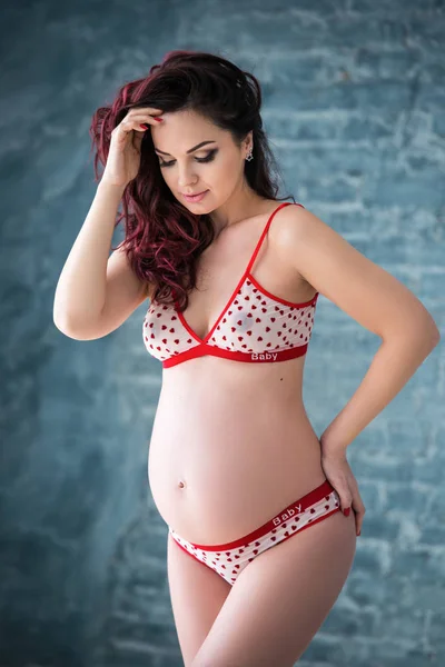 Mooie zwangere meisje vrouw in lingerie met een patroon van harten staat tegen een grijze bakstenen muur. Gezond moederschap en gelukkige zwangerschap — Stockfoto