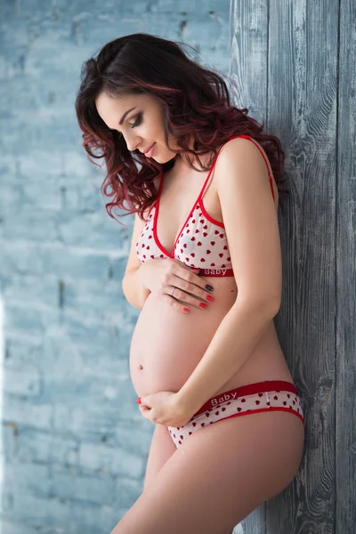 Beautiful pregnant girl woman in lingerie with a pattern of hearts is standing against a gray brick wall. Healthy motherhood and happy pregnancy — Stock Photo, Image