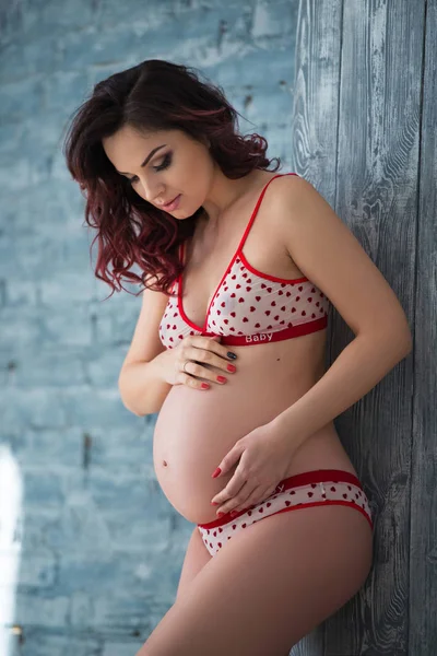 Bela mulher grávida em lingerie com um padrão de corações está em pé contra uma parede de tijolo cinza. Maternidade saudável e gravidez feliz — Fotografia de Stock