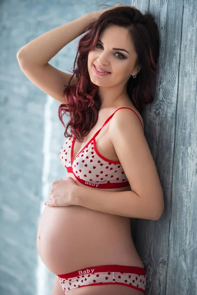 Vacker gravid tjej kvinna i underkläder med ett mönster av hjärtan står mot en grå tegelvägg. Friska moderskap och Happy graviditet — Stockfoto