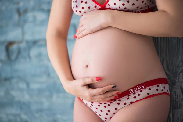 Těsné břicho těhotné dívky v prádle se srdcem na šedé cihlové stěně — Stock fotografie